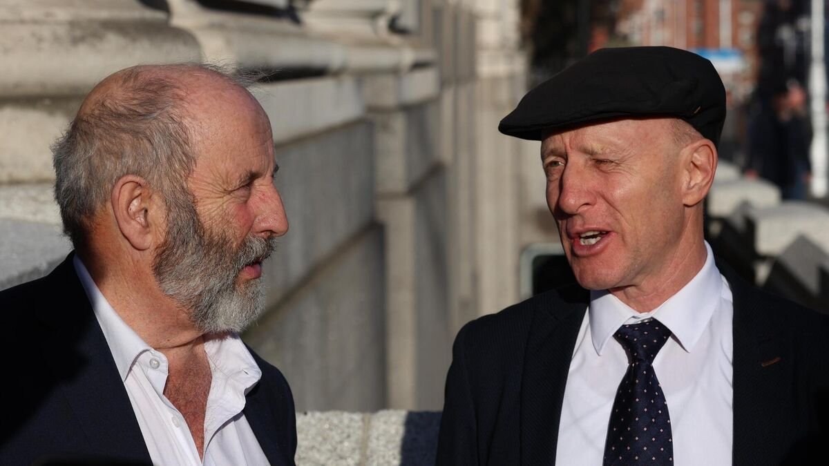 Michael and Danny Healy-Rae, two Irish Independent TDs, engaged in conversation outside a government building. Michael Healy-Rae is wearing a dark flat cap and suit, while Danny Healy-Rae is dressed in a suit with an open-collar shirt.